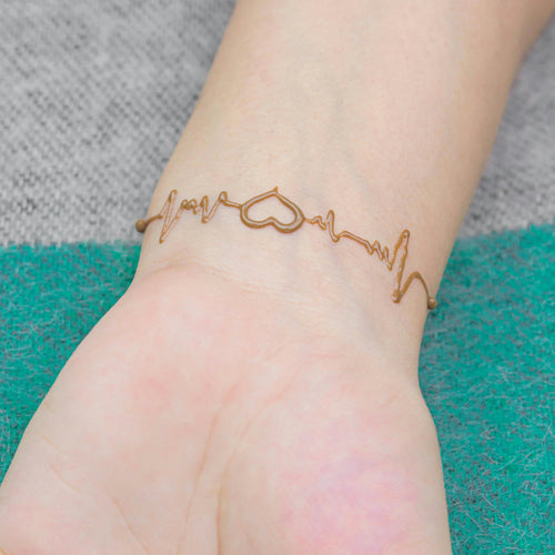 A person's wrist wearing a gold-colored bracelet designed to resemble a pulse waveform, placed on a green and gray fabric.